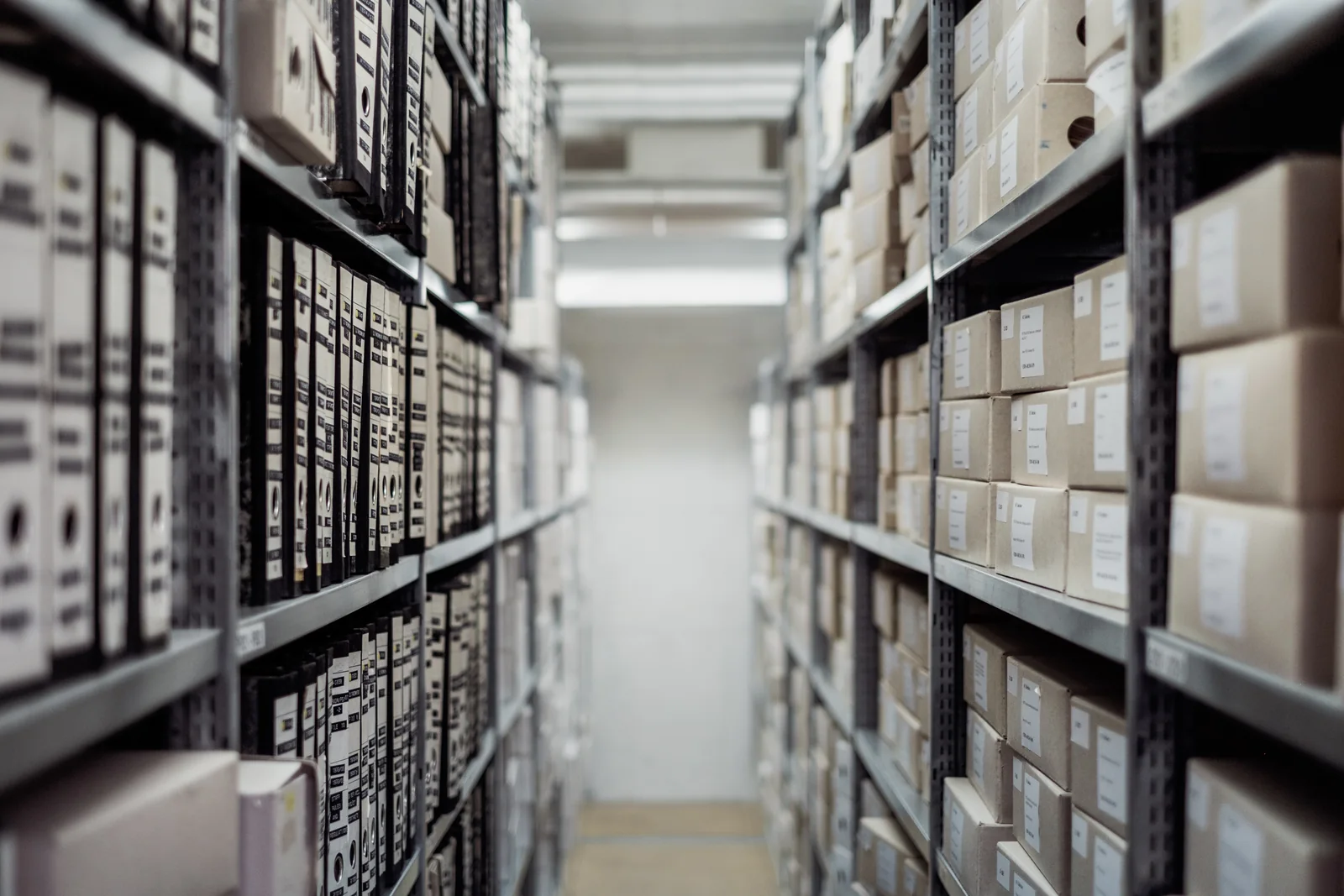 Two rows of grey metal shelving, lined with archive boxes and box files, run down a narrow aisle towards a pale wall.