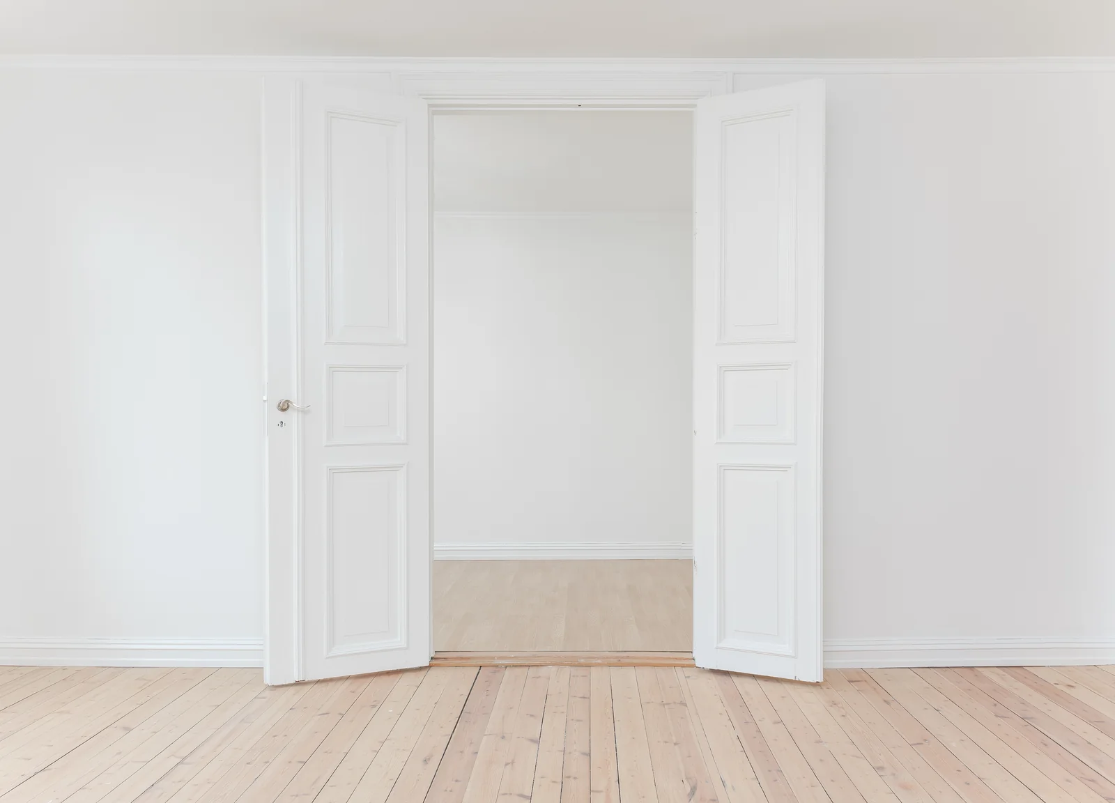 Open white double doors lead into a bright, empty room with a pale wooden floor.