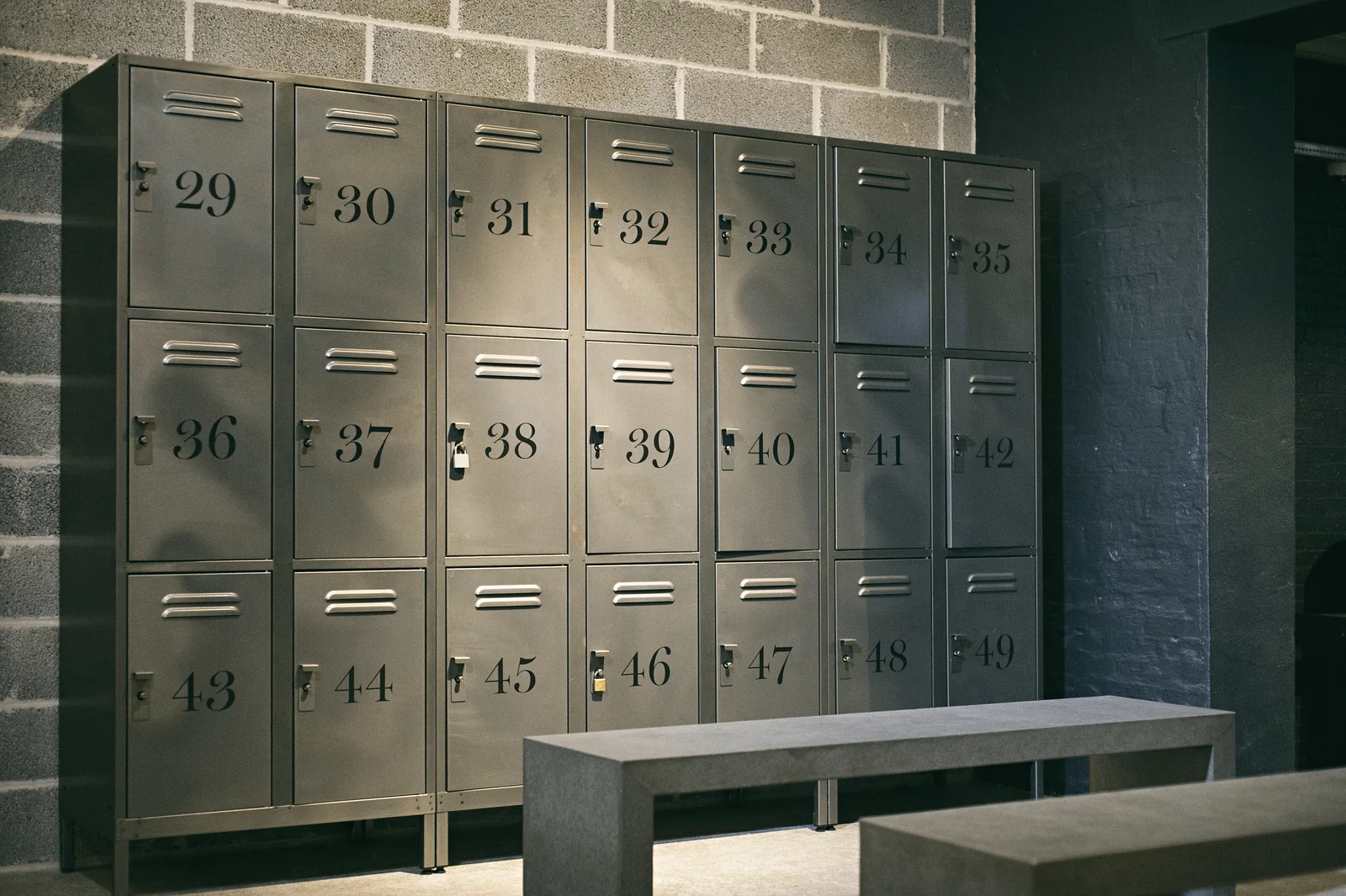 A bank of numbered metal lockers stands against a concrete block wall, with a concrete bench in front.