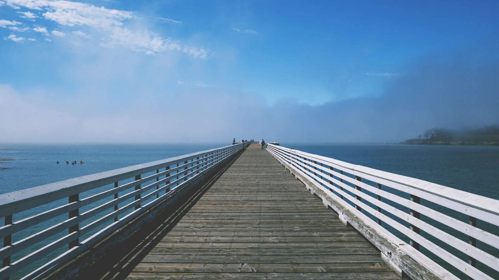 A long wooden pier with white railings runs out over calm blue water into haze.