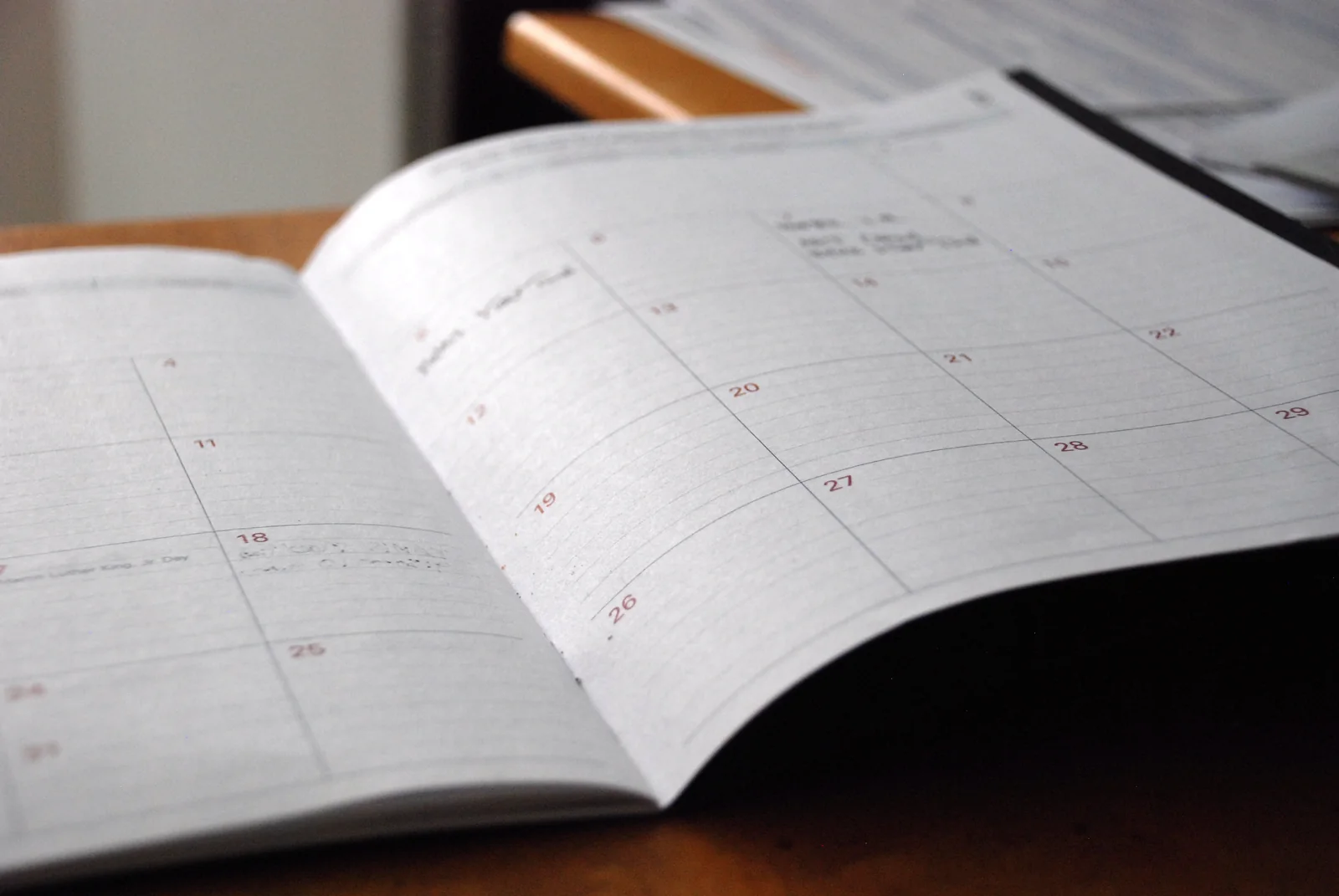 An open month planner lies on a wooden desk, its numbered squares mostly blank, against a dark background.