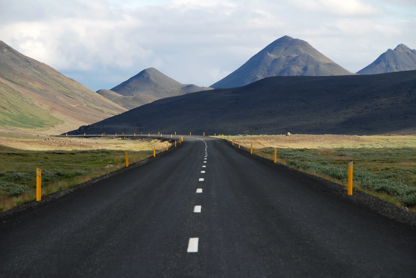 A straight road runs towards dark hills under a cloudy sky, with yellow marker posts along the verge.